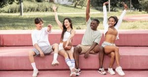 delighted multiethnic friends raising arms happily on bench in park